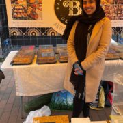Naz, of localcompany called Baklavalicious. Standing in gazebo market stall with trays of baklava.