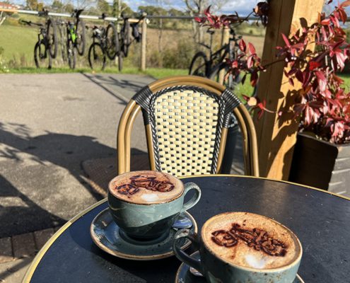 Two cappuccinos with a cocoa powder decoration of cycles with view of bikes and countryside behind