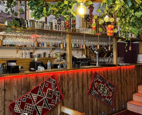 Photo of bar in Lebanese village Milddle Eastern restaauant, whith shelves of bottles, ethnic fabric hangings in front of the bar in warm lighting, and foliage decorating the bar above