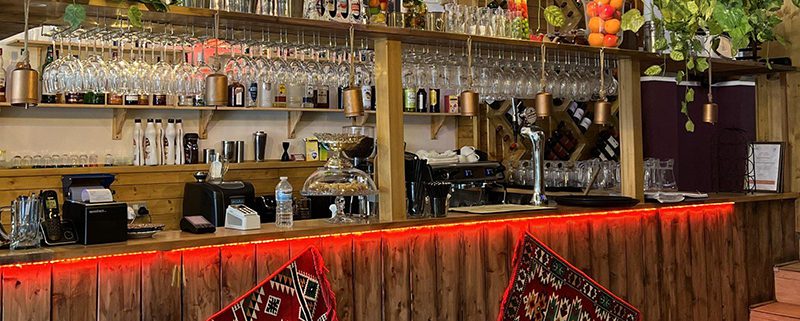 Photo of bar in Lebanese village Milddle Eastern restaauant, whith shelves of bottles, ethnic fabric hangings in front of the bar in warm lighting, and foliage decorating the bar above